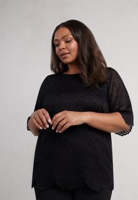 Top in pizzo nero con maniche corte, caratterizzato da un orlo scallop e un motivo intricato. Tessuto morbido e design dalla vestibilità rilassata.