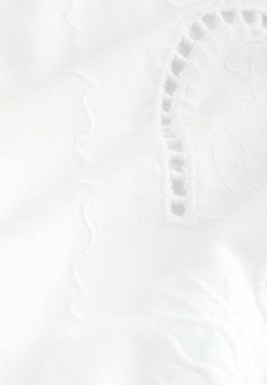 Valge kangas peene lillemustri tikandi ja dekoratiivsete väljalõigete detailidega, mis näitavad tekstuuri ja õrnu kõrgendatud mustreid.