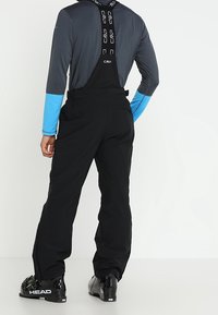 Zwarte skibroek met verstelbare banden, gecombineerd met een grijze langarmshirt met blauwe mouwen. Gekenmerkt door zwarte skischoenen.