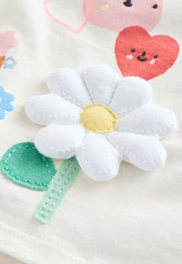 White flower applique with a yellow center on a cream fabric, accompanied by a green leaf. Colorful cartoon hearts and flowers in the background.