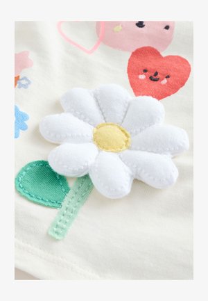 Fehér virágapplikáció sárga középpel krémszínű szöveten, zöld levéllel kísérve. Színes rajzfilm stílusú szívek és virágok a háttérben.