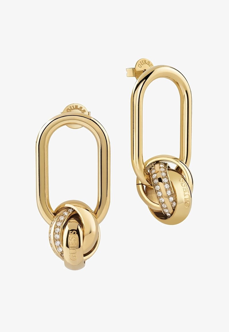 Boucles d’oreilles créoles dorées avec une forme ovale allongée et deux anneaux entrelacés, dont un orné de petits cristaux et gravé du logo GUESS.
