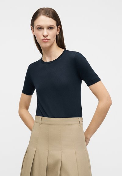 Γυναίκα με ίσια καστανά μαλλιά φοράει μπλε σκούρο μπλουζάκι με κοντά μανίκια, το οποίο είναι μαζεμένο σε μια υψηλοκάβαλη μπεζ πλισέ φούστα, μπροστά σε ένα απλό φόντο.