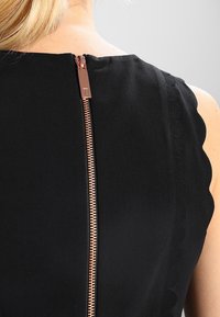 Robe noire avec un décolleté festonné et une fermeture éclair en or rose au dos, mettant en valeur une texture de tissu lisse.