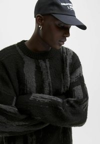 Junge Person trägt einen schwarz-grau gemusterten, flauschigen Pullover, eine schwarze Mütze mit weißem Text, silberne Creolen und Ohrstecker, schaut nach unten vor einem weißen Hintergrund.