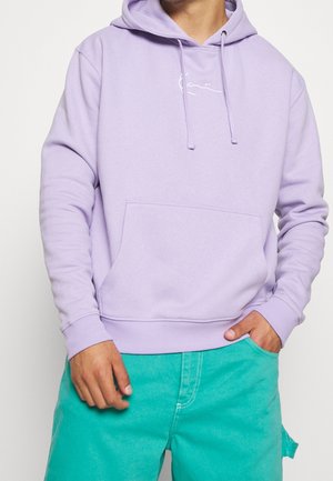 Mann trägt einen pastell-lila Hoodie mit Fronttasche und türkisfarbenen Shorts, steht vor einem einfarbigen hellen Hintergrund.