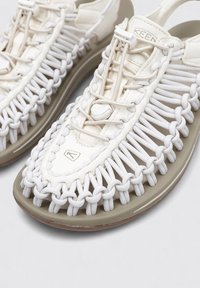 Sandalias deportivas con una suela de goma beige y una parte superior blanca texturizada que presenta un intrincado diseño trenzado y un sistema de cordones ajustable.