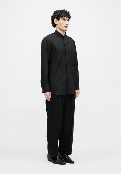 Melna garroku krekls ar stāvošu apkakli un kvadrātveida kabatas detaļu. Kombinēts ar brīvām melnām biksēm un melnām smailām kurpēm.