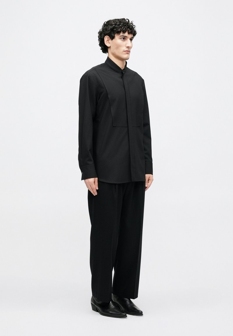 Schwarzes Langarmhemd mit Stehkragen und einer quadratischen Taschenapplikation. Kombiniert mit locker sitzenden schwarzen Hosen und spitzen schwarzen Schuhen.