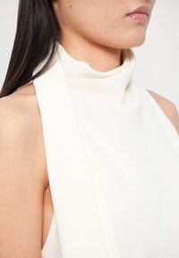 Balts bezroku tops ar augstu apkakli. Tam ir maiga, gluda tekstūra un drapējuma detaļa apkaklē.