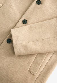Beige frakke med et blødt, tekstureret stof, prydet med sorte knapper og en glat krave. Inkluderer sidelommer og en struktureret design.
