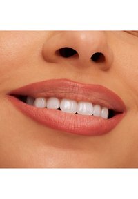 Nærbilde av en smilende munn med ferskenfarget matt leppestift, hvite tenner og glatt hudtekstur. Naturlige leppekonturer synlige.