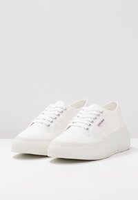 Białe płócienne sneakersy z teksturowaną podeszwą, z metalowymi oczkami, płaskimi sznurowadłami i detalem logo z boku. Okrągły kształt noska.