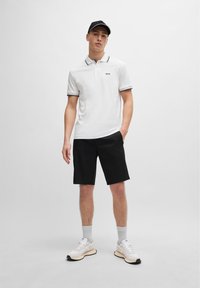 Cămașă polo albă cu guler și margini ale manșetelor în contrast negru, combinată cu pantaloni scurți negri, adidași albi și o șapcă neagră. Textură netedă.