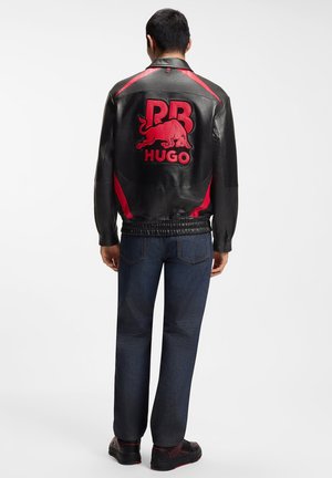 Man draagt een zwarte leren jas met het Red Bull- en "PB HUGO"-logo op de rug, donkere spijkerbroek en zwarte schoenen met rode accenten, staat stil.