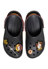 Zuecos negros con pins decorativos de temática Gryffindor, que incluyen un león, una espada y el emblema de la casa; superficie texturizada con orificios de ventilación.