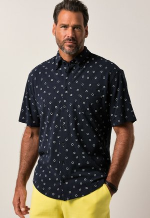 Man met baard die een zwart overhemd met korte mouwen met witte patronen en felgele broek draagt, staand met één hand in de zak.