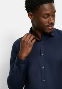 Marineblaues Button-up-Hemd aus strukturiertem Stoff, mit spitzem Kragen und langen Ärmeln; hat kontrastierende Knöpfe an der Knopfleiste und den Manschetten.