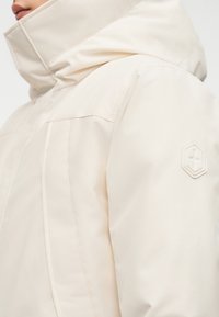 Αδιάβροχο μπεζ ανοιχτόχρωμο μπουφάν με ψηλό γιακά, από υφασμάτινη υφή, με πλευρικές τσέπες και εξαγωνικό λογότυπο στο μανίκι.
