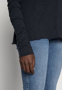 Camisa de manga longa, cinza escuro, com um corte descontraído e bainha sem acabamentos, combinada com jeans ajustados de azul claro. A mão repousa na coxa.