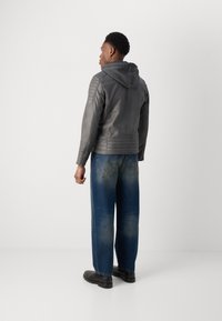 Graue Lederjacke mit Kapuze, ausgestattet mit gepolsterten Schulterdetails und taillierter Passform, kombiniert mit ausgewaschenen blauen Weitbeinhosen und schwarzen Schuhen.