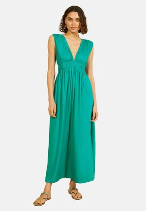 Kvinde iført ærmeløs tealgrøn maxikjole med dyb V-halsudskæring og flade sandaler, stående mod ensfarvet hvid baggrund.