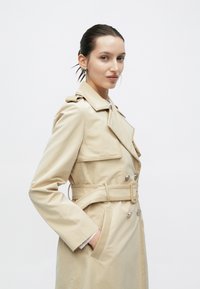 Μπεζ παλτό τρεντς με ζώνη στη μέση, επιχρυσωμένες ωμοπλάτες, διπλό κούμπωμα και πλαϊνές τσέπες· λείο, αδιάβροχο ύφασμα.