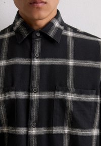 Chemise en flanelle à carreaux noir et blanc avec un col, deux poches avant et fermeture à boutons. Tissu texturé avec des lignes blanches contrastantes.