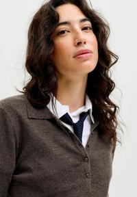 Mlada žena s dugom kovrčavom tamnom kosom, koja nosi smeđi duksericu sa dugmadi, bijelu košulju s kragnom i plavu prugastu kravatu, koja gleda malo prema gore.