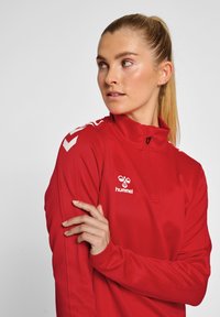 Veste de sport rouge à demi-zip en tissu lisse, avec des détails blancs et un logo sur la poitrine gauche. Manches longues et design ajusté.