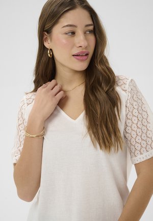 Jeune femme aux cheveux longs et ondulés portant un haut blanc à col en V avec des manches en dentelle, des boucles d'oreilles en or en forme d'anneau, un bracelet, des bagues et un collier délicat, regardant vers la droite.