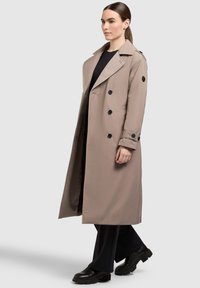 Beige trenchcoat met een dubbelzijdig ontwerp, zwarte knopen en een brede kraag; gedragen over zwarte kleding en gecombineerd met stevige zwarte schoenen.