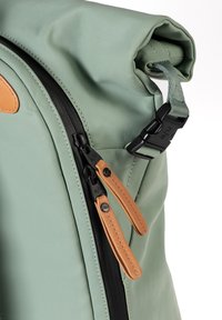 Zaino roll-top in tessuto verde tenue con zip nera, dettagli in pelle e tracolla regolabile. Caratterizzato da una superficie liscia e un design minimalista.