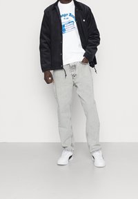 Giacca di velluto a coste nera sopra una felpa bianca con grafiche, jeans grigio chiaro e sneakers bianche con accenti neri. Outfit casual.