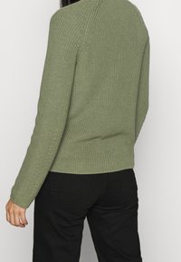 Gebreide trui in lichtgroen met lange mouwen, geribbelde textuur en zijslitsen. Gecombineerd met zwarte broek, gezien van achteren.