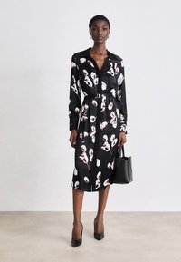 Černé květinové šaty s dlouhými rukávy, límcem, pásečkem v pase a ke kolenům dlouhým lemem. Nošeny s černými lodičkami a malou černou kabelkou.