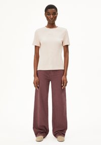 Lichtbeige geribbelde top met korte mouwen, gecombineerd met wijde, diep bordeauxrode broek en lichtbeige schoenen, voor een effen witte achtergrond.