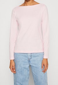 Helles pinkes Langarmoberteil mit weitem Ausschnitt, aus weicher Baumwolle. Es hat einen geraden Saum und dezente Stickdetails. Getragen mit Jeans.