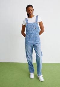 Denim overaller i lys blå med justerbare stropper, lomme foran og løs passform, kombinert med en hvit T-skjorte og hvite joggesko.