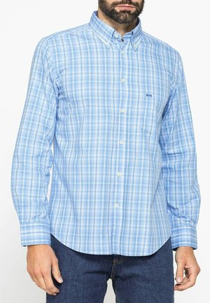 Uomo che indossa una camicia a quadri azzurri chiari con bottoni e maniche lunghe e jeans blu scuro, in piedi davanti a uno sfondo bianco.