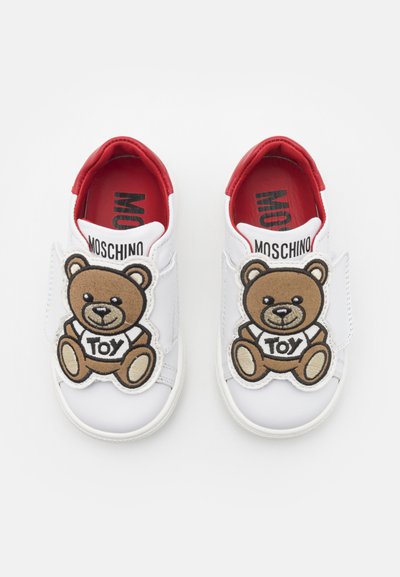 Hvite skinnsneakers med rødt innvendig. Har et stort brunt bjørne-patch på borrelåsen, som har på seg en "TOY"-t-skjorte. Flat gummisåle.