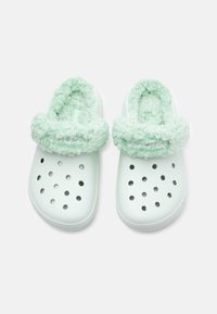 Sabots blancs à enfiler avec de ronds trous de ventilation et un doux rembourrage en fourrure verte claire, présentés de haut sur un fond blanc.