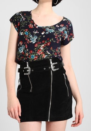 Femme portant un top court à manches courtes bleu marine à motif floral, rentré dans une mini-jupe noire avec des détails de fermeture éclair argentés et une ceinture à double boucle.