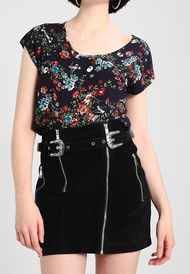 Femme portant un top court à manches courtes bleu marine à motif floral, rentré dans une mini-jupe noire avec des détails de fermeture éclair argentés et une ceinture à double boucle.