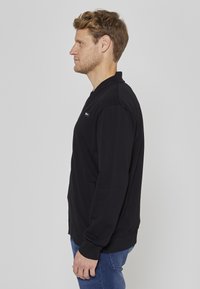 Sweatshirt noir en coton, avec un col rond, des poches latérales et des poignets côtelés, présenté de côté sur un fond uni.