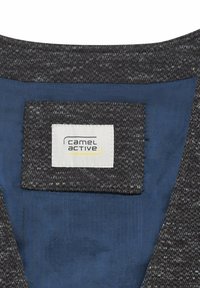 Cuello de chaqueta de tela texturizada gris oscuro con forro azul y una etiqueta rectangular blanca que dice "camel active" en texto negro y amarillo.