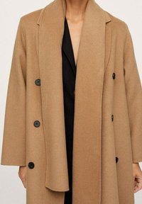Manteau en mélange de laine couleur chameau avec un design à double boutonnage, de gros boutons noirs et de larges revers. Présente une coupe décontractée et des manches longues.