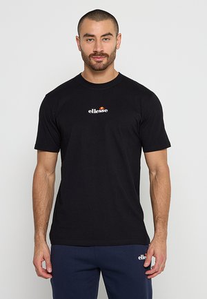 Hombre con cabello corto y barba, vistiendo camiseta negra ellesse y pantalones azul marino ellesse, de pie frente a un fondo claro y simple.