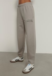 Šedé tepláky s elastickým pásom, bočnými vreckami a vyšívaným logom "LAMANIA" v čiernej farbe. Skombinované s bielymi a čiernymi teniskami.