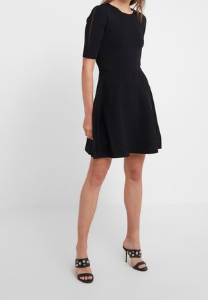 Robe noire à manches courtes avec un corsage ajusté et une jupe évasée, présentant un ourlet festonné. Associée à des sandales à talons noires à brides.